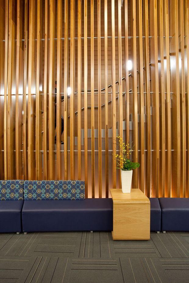 A contemporary commercial reception or waiting area featuring a long dark navy upholstered bench, a patterned backrest, and a light timber side table with a potted plant. The backdrop is a dramatic full-height screen of closely spaced vertical timber battens, with a staircase and warm lighting visible behind it. Grey patterned carpet tiles cover the floor.