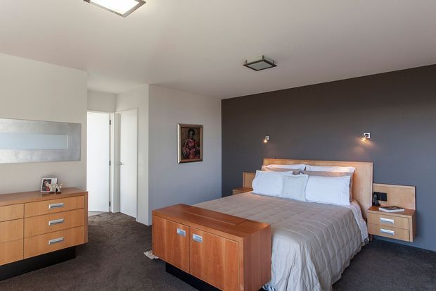 A contemporary bedroom with a large upholstered bed against a dark grey feature wall, warm timber bedside tables, and a matching timber storage bench at the foot of the bed. The room has dark carpet, white walls and ceiling, recessed and square ceiling lights, small wall-mounted bedside lights, and a timber drawer dresser. A doorway and framed artwork are also visible.