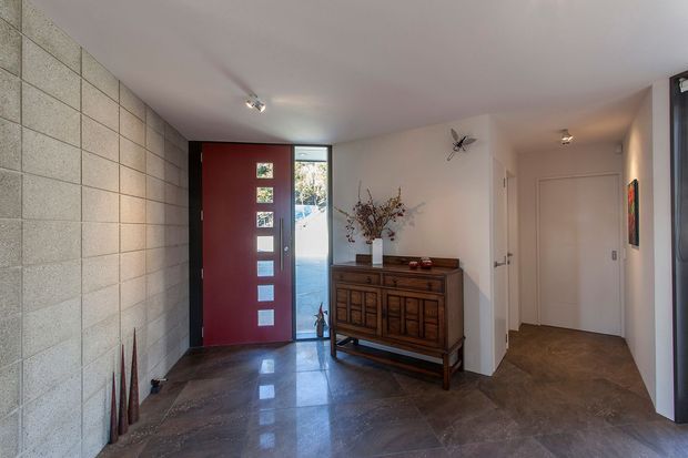Interior view of a modern residential entry hall featuring a tall deep-red front door with vertical glazing, an exposed concrete-block feature wall, polished dark stone-look flooring, white walls and ceiling, and a vintage timber sideboard with decorative objects. A short hallway leads to additional rooms, with daylight entering through the front door glazing.