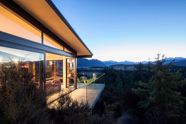 A modern single-storey home viewed from an elevated outdoor deck at dusk, with expansive floor-to-ceiling glazing, dark metal window frames, and a broad projecting roof eave. Warm interior lighting reveals a living area inside, while the deck overlooks a forested landscape and layered mountain ranges. The composition emphasizes the building’s connection to its scenic natural setting.