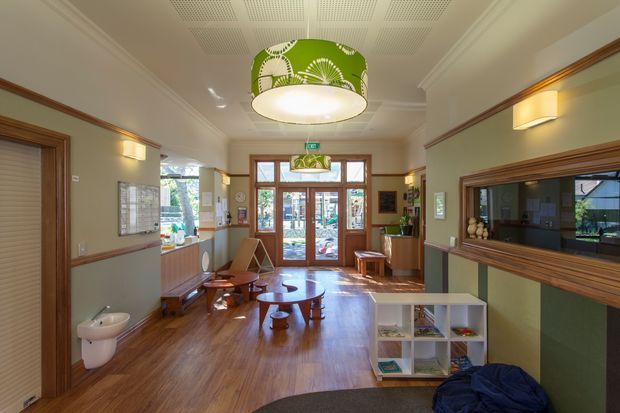 A warmly lit reception or entry lobby with polished timber flooring, wood-framed glazed entry doors, timber trim and panelling, pale walls, and a large interior service or viewing window. The space includes lounge seating, a low coffee table, shelving, wall sconces, and a decorative green pendant light. A partially visible kitchenette or service area opens to the left, while the overall room has a welcoming, heritage-inspired fit-out.