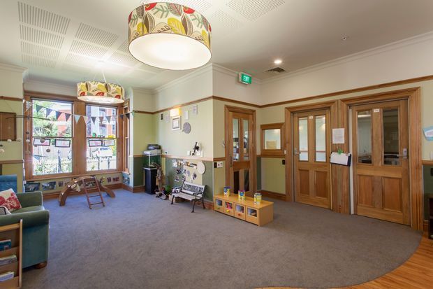 A spacious institutional interior, likely a school or community facility lobby, with pale green walls, extensive warm timber doors and window frames, carpeted flooring, and a patterned cylindrical pendant light. The room includes child-oriented seating, a low colorful storage unit, noticeboards, artwork, and glazed timber doors leading to adjoining spaces. Decorative acoustic ceiling panels and traditional trim details are visible.
