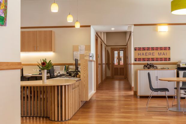 A warm, welcoming reception and waiting area with a timber slatted reception desk, timber cabinetry, polished wood flooring, pendant lighting, and a corridor leading deeper into the building. A Māori “Haere Mai” welcome sign and patterned artwork add cultural character, while a small table and chairs form a waiting area on the right.