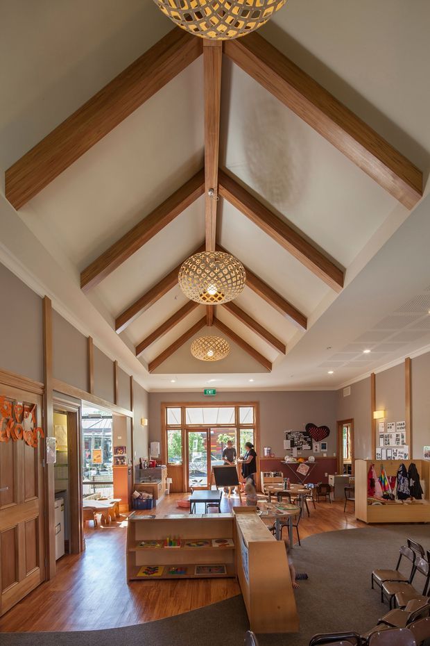 Interior view of a welcoming educational or community space with a long, high vaulted ceiling supported by exposed warm timber rafters and beams. Multiple woven pendant lights hang along the central ridge. The room has timber flooring, warm taupe walls, timber-framed glazing and doors, display boards, low shelving, tables and chairs, and an entrance at the far end. The space appears to function as a classroom, library, reception, or community learning area.
