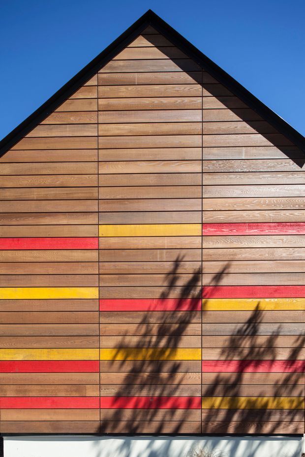 Close-up exterior view of a steep triangular gable clad in horizontal timber boards. The warm brown wood facade is accented with scattered horizontal bands of bright red and yellow, framed by dark roof trim. Strong sunlight casts branch shadows across the lower facade, with a clear blue sky behind.