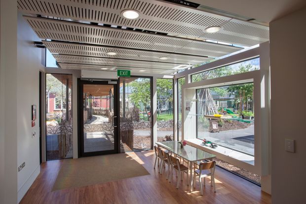 A bright, contemporary communal dining or breakout area with a long glazed wall and double glass entry doors opening to an outdoor courtyard or playground. The room has warm timber-look flooring, white walls, a suspended linear slatted ceiling with integrated circular downlights and ventilation grilles, and a compact timber dining table with chairs. An exit sign and fire equipment indicate an institutional or commercial setting.
