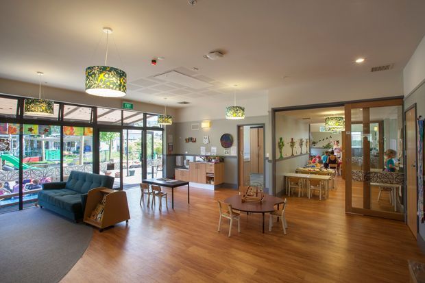 A bright, open interior of a community or childcare facility with a lounge and reception area in the foreground, a dining or activity room visible through large timber-framed glazed doors, and extensive glazing along the left side. The space features warm timber-look flooring, colourful patterned pendant lights, exposed services in the ceiling, built-in cabinetry, child-sized tables and chairs, and a playful, welcoming atmosphere.
