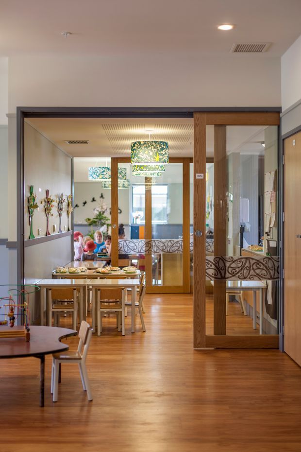 A bright, warm interior view of a childcare or educational dining and activity area. The space features timber-look flooring, wood-framed glazed sliding doors with decorative patterned panels, mirrored or reflective glazing, child-sized tables and chairs, display shelving, and colourful artwork and pendant lighting. The view looks through a series of connected rooms toward a glazed exterior opening.