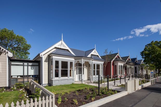 Street-facing view of a row of attached heritage-style cottages with ornate timber gables, decorative fretwork, bay windows, contrasting painted weatherboard façades, and small landscaped front gardens. A paved accessible walkway and low picket fencing run along the frontage under a clear blue sky.