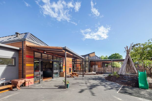 Exterior view of a low-rise contemporary building arranged around a paved courtyard. The facade combines dark metal cladding with warm horizontal timber weatherboards and broad glazed entrances, beneath angular standing-seam metal roofs. The courtyard includes covered outdoor circulation, timber seating, fencing, mature planting, and a distinctive teepee-like timber play structure.