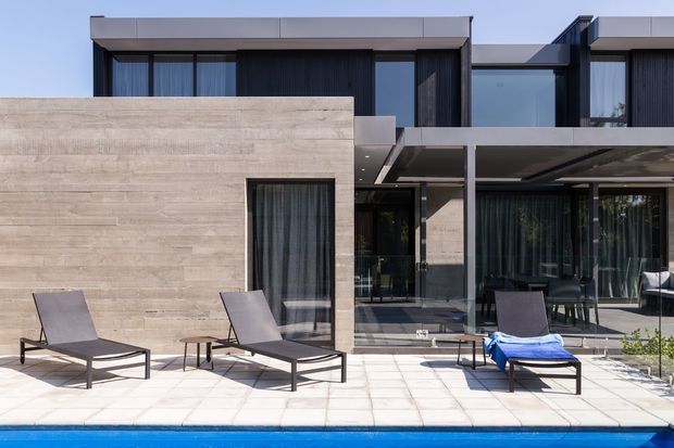 Exterior view of a contemporary two-storey residence beside a swimming pool. The home combines pale horizontal concrete or stone-look wall cladding with dark vertical cladding, extensive glazed openings, and a flat roof. A covered outdoor terrace with dining furniture and poolside sun loungers occupies the foreground.
