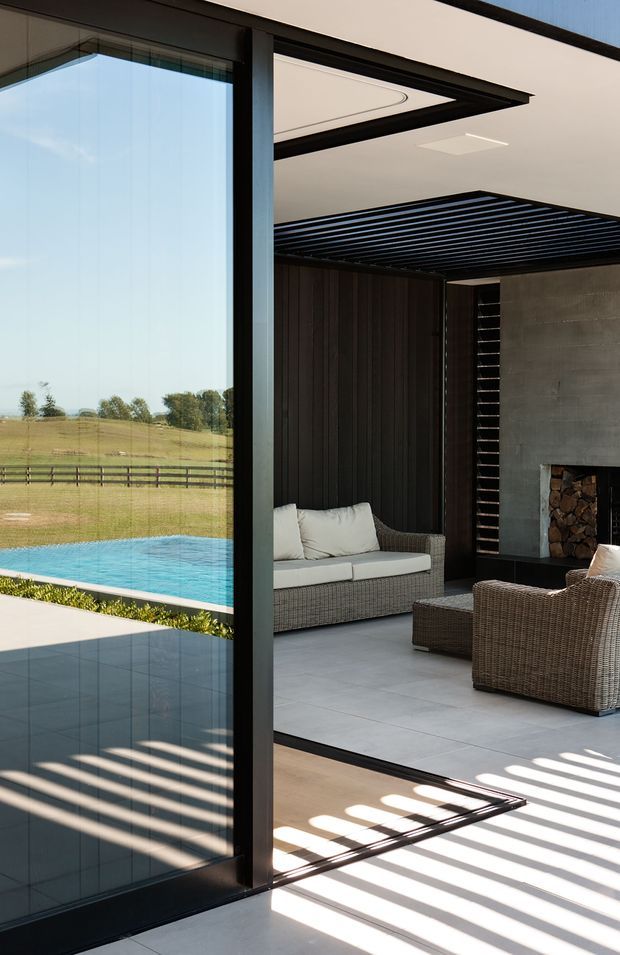 A contemporary indoor-outdoor living area viewed through a large black-framed glazed opening. The covered lounge features wicker seating with pale cushions, large light-grey tiled flooring, dark vertical wall cladding, a concrete fireplace with stacked firewood, and a dramatic black linear slatted ceiling detail. Beyond the glazing is a bright blue swimming pool and rural landscaped setting. Strong striped shadows fall across the floor from the overhead screening.