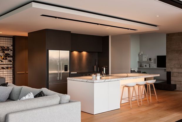 Open-plan contemporary kitchen and living area with a large white island and integrated seating, dark timber cabinetry, stainless-steel refrigerator, pale stone benchtop, and warm timber flooring. A grey sectional sofa occupies the foreground, while recessed linear lighting, display shelving, and a television define the adjacent living zone.