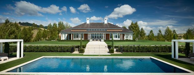 A symmetrical single-storey residence viewed from across a rectangular swimming pool. The house has a low hipped roof with two white chimney stacks, a long covered veranda supported by slender columns, and extensive glazed doors and windows. Formal clipped hedges, broad lawns, white-framed poolside structures, and a central stair approach create a carefully landscaped, balanced composition. Rolling green hills and trees are visible beyond the property under a bright blue sky.