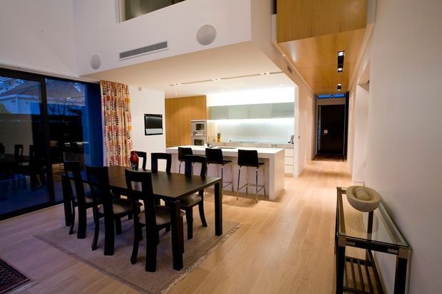 Open-plan contemporary interior combining a dining area, kitchen and hallway. A dark rectangular dining table with matching black chairs sits on a rug beside full-height glazing and patterned curtains. The bright white kitchen has a central island with bar seating, pale timber cabinetry and light timber flooring. Recessed lighting, integrated appliances, and a warm timber ceiling detail create a clean, refined space.