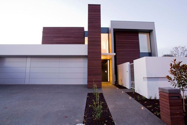 Contemporary multi-level residential facade viewed from the driveway, featuring clean white rendered volumes, a large sectional garage door, dark horizontal timber cladding accents, a tall vertical timber-clad architectural element, and a recessed illuminated entry along a paved walkway. Minimal landscaped garden beds and a low boundary wall frame the approach.