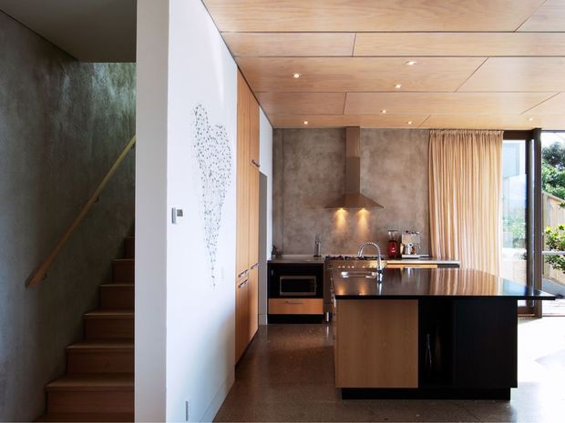 Compact contemporary kitchen viewed from an adjoining stair area, featuring a central island with stainless-steel sink and integrated appliances, dark stone-like benchtops, warm timber cabinetry, a timber-lined ceiling, and a textured concrete-toned splashback wall. Full-height glazing and curtains open the kitchen toward the outdoors, while a timber stair with a dark textured wall is visible at left.