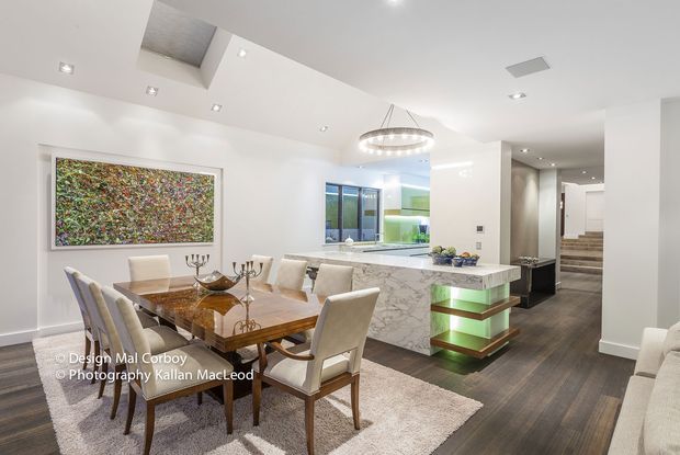 Bright open-plan dining and kitchen interior with a contemporary, minimalist fit-out. A polished marble island with illuminated open shelving separates the kitchen from the dining area, which features a timber dining table, upholstered chairs, a large neutral rug and a linear botanical artwork. Recessed ceiling lighting, a circular pendant, skylights and a stair hall contribute to the airy, refined space.