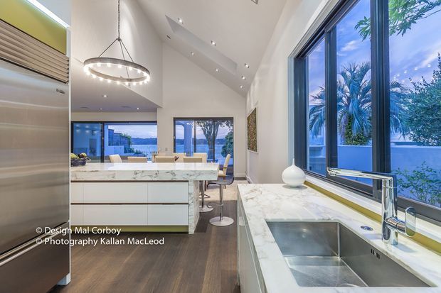 Bright contemporary kitchen with a large white marble-look island, integrated cabinetry, stainless-steel appliances, timber flooring, and a broad double-height glazed opening overlooking lush vegetation and water. A long marble-look benchtop with undermount sink runs beneath expansive windows, while a sculptural circular pendant light and recessed ceiling lights illuminate the space.
