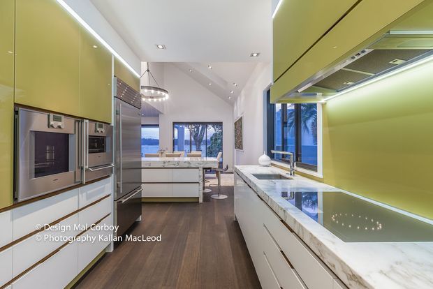 Contemporary galley kitchen with glossy lime-green full-height cabinetry, integrated stainless-steel appliances, white lower cabinets, and a white marble-look island and benchtop. A long island with seating sits beneath pendant lighting, while large windows at the far end bring in daylight and views of greenery and water. Dark timber flooring contrasts with the light cabinetry and bright finishes.