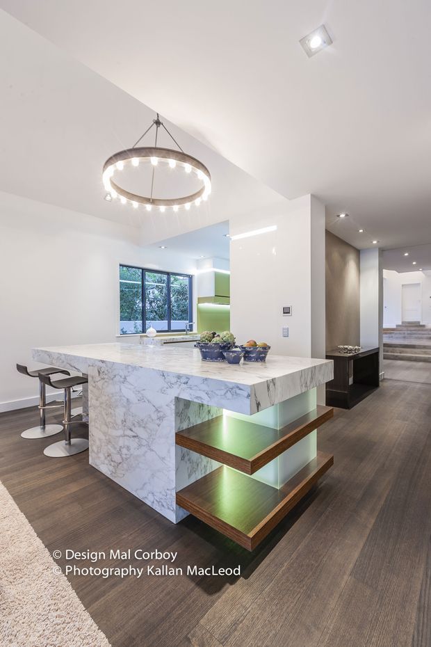 Bright contemporary kitchen interior dominated by a large white marble island with waterfall sides and open illuminated timber display shelves. The island includes two bar stools, while white cabinetry, integrated appliances, recessed lighting and a large pendant ring light create a clean, refined fit-out. Dark timber-look flooring contrasts with the white surfaces, and a hallway with stairs is visible beyond.