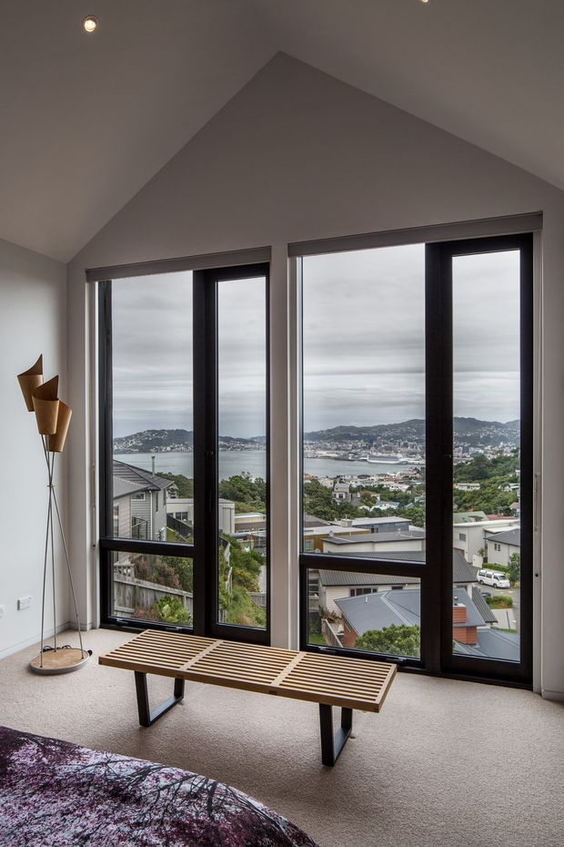 A contemporary bedroom with a high pitched ceiling and three tall black-framed windows overlooking a hilly coastal harbour settlement. The room has light neutral walls, beige carpet, a slatted timber bench at the windows, a sculptural floor lamp, and a partially visible patterned bedspread. The expansive glazing provides a strong connection to the surrounding landscape.