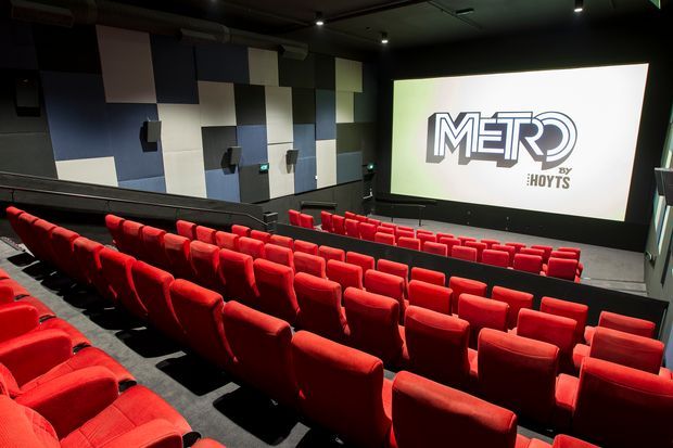 Interior view of a commercial cinema auditorium with multiple rows of bright red upholstered theater seats facing a large projection screen. The room has dark flooring and ceiling, integrated wall speakers, acoustic wall panels in a geometric pattern, and a prominent Metro by Hoyts logo on the screen.