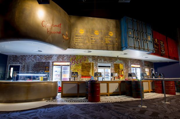 A dark, industrial-style commercial food and beverage venue with a long service counter, illuminated display fridges, menu boards, exposed red brick walls, and distinctive shipping-container-inspired blue and red wall features. Curved metallic ceiling soffits and barrel tables add a modern hospitality fit-out, while patterned carpet and queue barriers occupy the foreground.