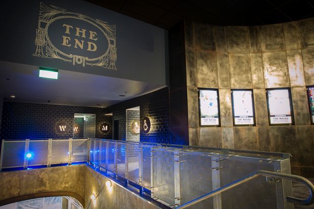 Dark, atmospheric interior of a cinema or entertainment venue with a projecting sign reading “THE END,” illuminated movie posters, black brick walls, metallic distressed wall panels, and a balcony or stair landing enclosed by perforated metal railings with blue accent lighting.