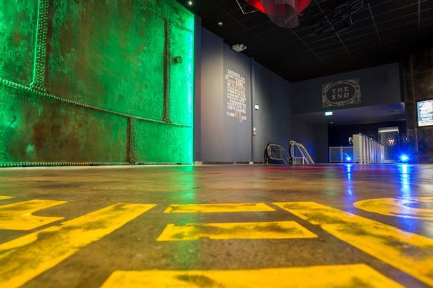 A dark, atmospheric interior of a themed entertainment or event venue, featuring a large weathered green metal feature wall, polished concrete floor with bold yellow graphics, black ceiling services, dramatic green, blue and red lighting, and signage including “The End.”