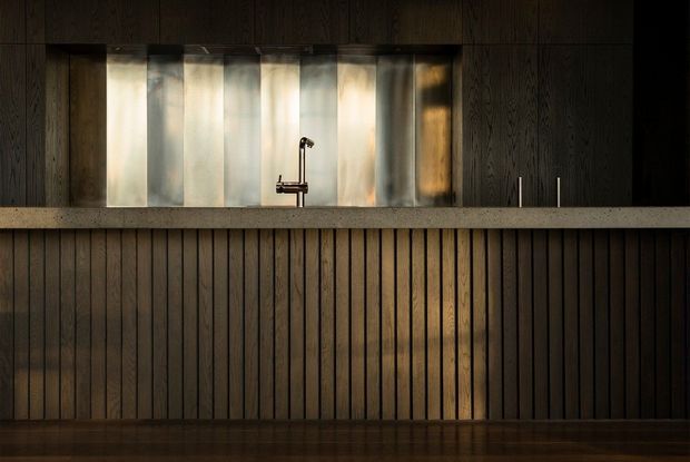 A dark, moody kitchen interior dominated by floor-to-ceiling vertical timber panelling and matching dark timber cabinetry. A long stone or concrete benchtop runs across the frame, with a compact metal tap and sink set beneath a softly diffused illuminated window or splashback. Minimal visible hardware and warm reflected lighting create a refined, intimate atmosphere.