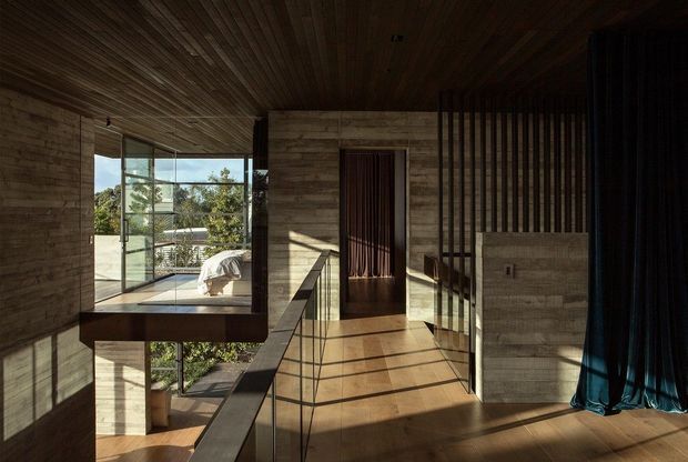 A dark, refined modern interior corridor or landing with extensive timber lining across the ceiling and walls, warm timber flooring, exposed structural or balustrade elements, and a dramatic double-height glazed opening. Sunlight enters from the left, revealing a lower-level garden view and an adjacent bedroom or living area beyond the glazing. Vertical timber screening and dark curtains add texture and privacy.
