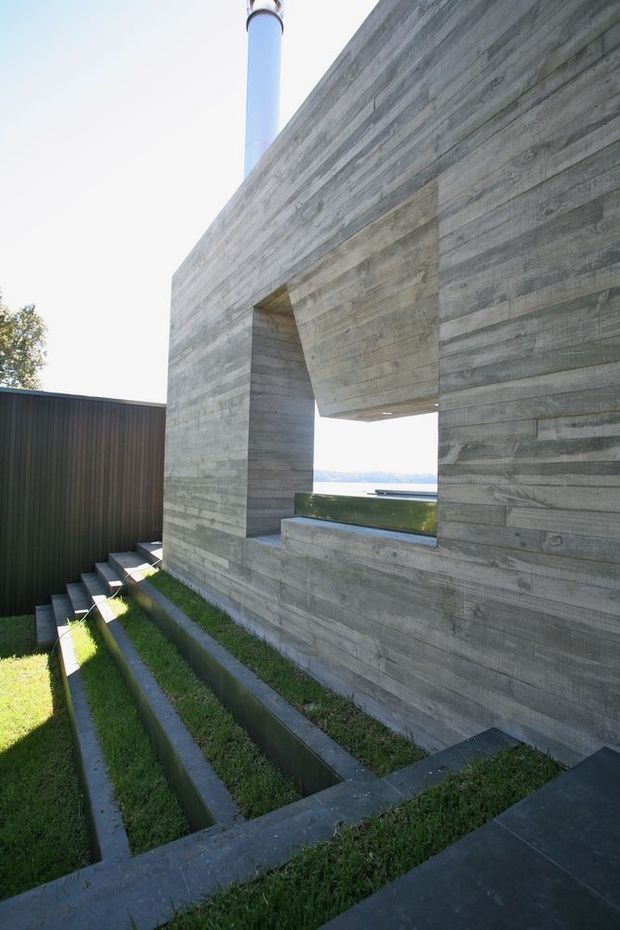 Exterior architectural view of a stark, rectilinear concrete structure with a large framed opening overlooking a distant body of water. The building has a rough horizontal board-formed concrete finish, dark vertical metal cladding, a cylindrical chimney or flue, and a series of narrow stepped grass channels and concrete-edged landscaping in the foreground.