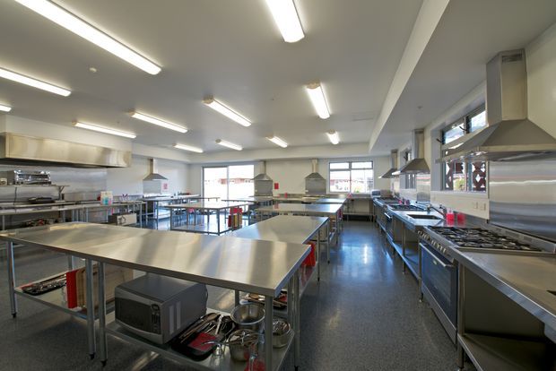 A large commercial teaching or professional kitchen with multiple stainless-steel worktables, cooking ranges, extraction hoods, sinks, shelving, and commercial appliances. The space has a practical open layout, polished dark flooring, white walls and ceiling, abundant fluorescent strip lighting, and windows along the rear and side walls.