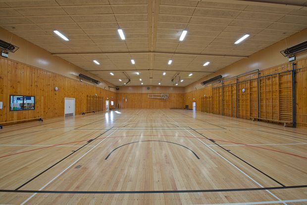 Large indoor sports hall or gymnasium with a polished timber multi-court floor, extensive timber wall panelling, wall-mounted climbing bars, high acoustic-panelled ceiling, and bright linear fluorescent lighting. The space is empty and designed for basketball, volleyball, and other court sports.