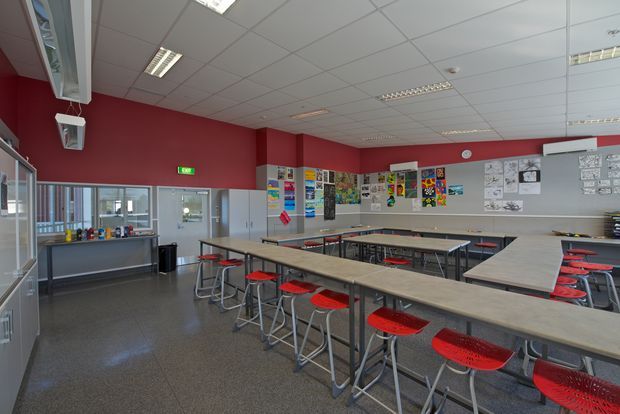 A spacious school art or technology classroom with rows of long light-coloured worktables and red stools. The room has deep red painted walls, grey polished flooring, suspended acoustic ceiling tiles with fluorescent strip lighting, extensive student artwork and posters on the walls, windows and a glazed door at the rear, and built-in shelving and display areas along the sides.