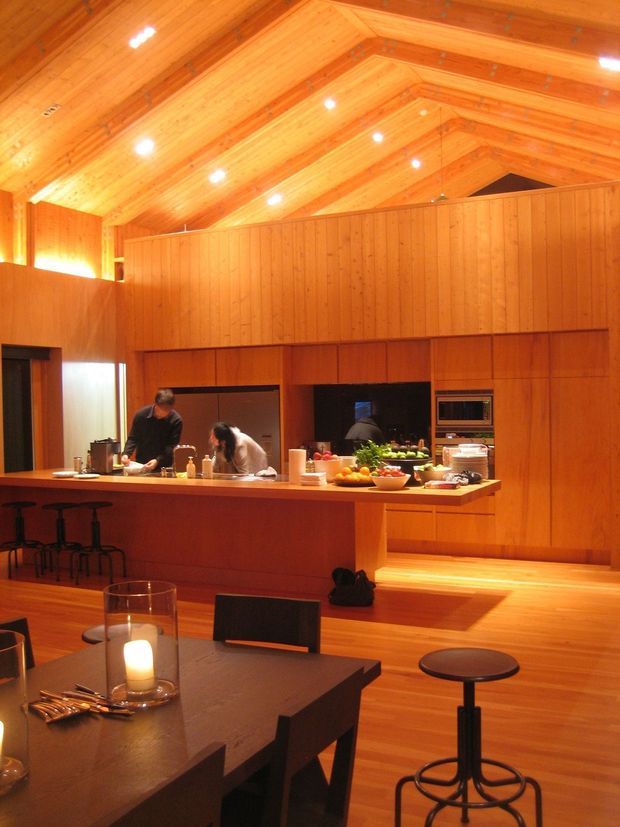 A warm, expansive open-plan kitchen and dining area dominated by extensive timber wall panelling and a dramatic pitched, timber-lined ceiling. The kitchen has a long island bench with stools, integrated appliances and open preparation space. A dining table occupies the foreground, while a partial upper-level mezzanine or balcony is visible above the kitchen. Recessed ceiling lights and warm timber surfaces create a cohesive amber-toned interior.