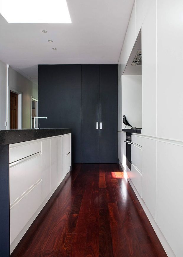 A narrow, contemporary galley kitchen with white full-height cabinetry, a dark charcoal pantry or storage wall, white lower cabinets, and a dark speckled stone benchtop. Rich dark reddish-brown timber flooring runs through the space. Recessed ceiling lights and a large rectangular skylight provide illumination, while integrated appliances and minimal hardware create a clean, streamlined appearance.