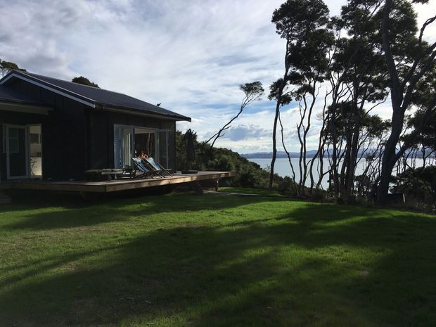 A dark-clad contemporary single-storey house with a low pitched roof and broad timber deck, set within a large grassy coastal landscape. Full-height glazed doors open onto the deck, where outdoor loungers are visible. Tall windswept trees frame a distant body of water and shoreline beyond.