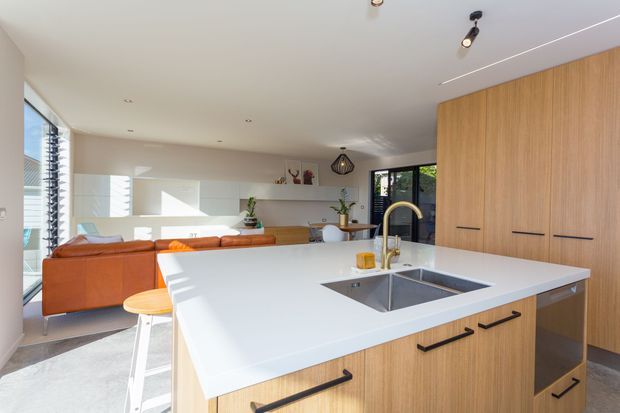 Bright open-plan kitchen and living area with a large white island, integrated double sink and curved brass tap, light timber cabinetry, and a warm cognac leather sofa. The space features expansive glazing, streamlined white walls, recessed lighting, and contemporary built-in joinery.