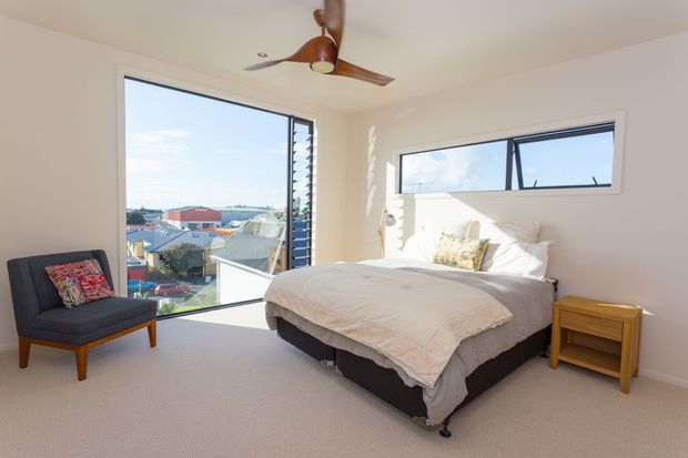 Bright contemporary bedroom with a large upholstered bed, light carpet, white walls, expansive corner glazing, a narrow horizontal window, timber ceiling fan, and a small timber bedside table. The windows overlook a suburban neighbourhood and water or coastline in the distance.