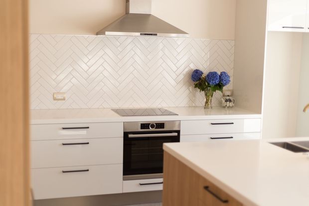Bright contemporary kitchen with white handleless cabinetry, a white engineered-stone benchtop, stainless-steel rangehood and appliances, and a white herringbone tile splashback. A timber-sided island with a pale benchtop occupies the foreground, while a vase of blue flowers adds colour.