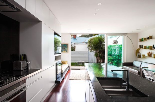 Bright contemporary open-plan kitchen and living area with sleek white full-height cabinetry, integrated appliances, a dark stone island benchtop with double sink, and polished dark timber flooring. The space opens through large glazed doors to a planted outdoor area, with a compact living zone and curved open shelving visible on the right.