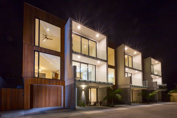 Night-time exterior view of a row of contemporary multi-storey townhouses. The buildings feature strong geometric forms, extensive floor-to-ceiling glazing, warm timber-clad facade sections, pale rendered walls, recessed balconies, and integrated exterior lighting. Ground-level garages and landscaped entry areas are visible.