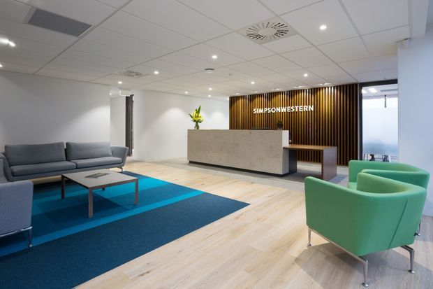 A bright contemporary commercial reception and waiting area with a pale timber-look floor, large blue geometric rug, grey sofa, green lounge chairs, and a stone reception desk. A black vertical timber-slat feature wall with illuminated signage forms the focal point, beneath a white suspended acoustic tile ceiling with recessed lighting.