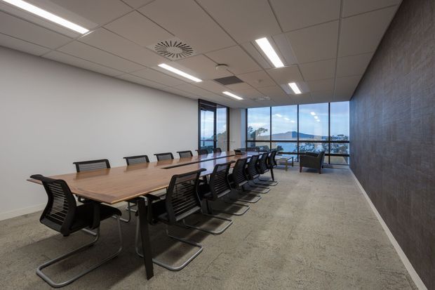 A contemporary commercial meeting or boardroom with a long timber conference table surrounded by black mesh cantilever chairs. The room has neutral painted walls, grey carpet, a suspended acoustic tile ceiling with recessed linear lighting, and a dark textured feature wall. Full-height glazing at the far end provides expansive views toward water and hills.