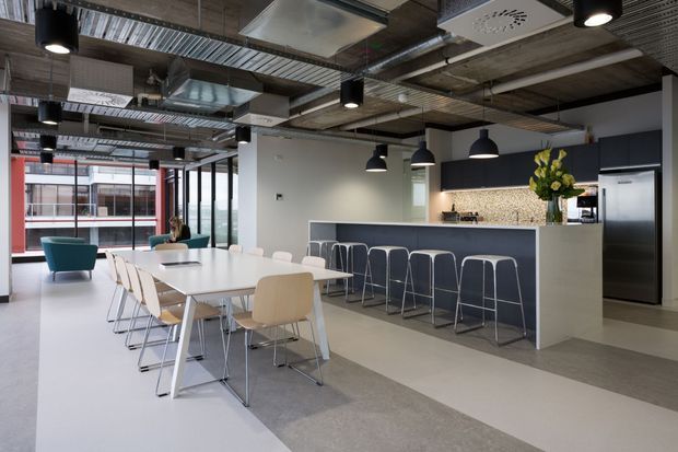 Contemporary open-plan commercial staff breakout and kitchenette area with a long dining table, bar-height island seating, exposed services and concrete ceiling, large glazed windows, and a restrained grey, white, and charcoal palette.