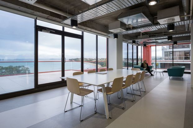 A contemporary open-plan commercial meeting and breakout space with a long white conference table, multiple chairs, teal lounge seating, exposed concrete ceilings, visible services, black-framed floor-to-ceiling glazing, and expansive waterfront and urban views. The interior has an industrial-influenced fit-out with bright natural light and a clean, spacious layout.