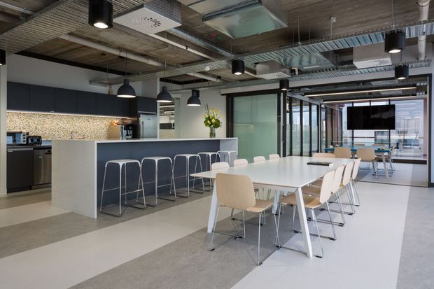 Open-plan contemporary office breakout and dining area with a communal white table, light chairs, kitchen island with bar stools, and glazed meeting or collaboration spaces beyond. The space features exposed services and ductwork, acoustic ceiling panels, black pendant lighting, dark kitchen cabinetry, a patterned backsplash, and a neutral polished concrete-style floor.