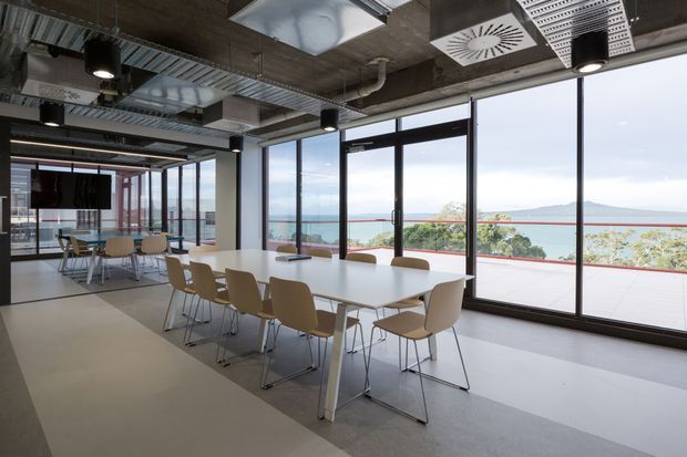 A contemporary commercial office dining and breakout area with a large communal white table and tan chairs beside full-height glazing. The space has an exposed concrete ceiling, visible services and ductwork, black pendant lights, linear air-conditioning grilles, and polished light flooring. Through the windows is a broad coastal or waterfront outlook with a terrace and distant hills.